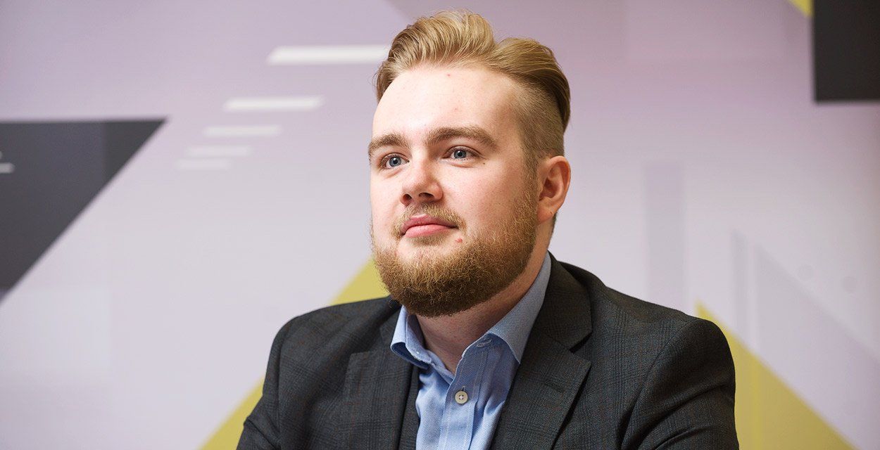 Man with a beard in a blazer, blue shirt. Sitting in an office setting, looking off-camera.