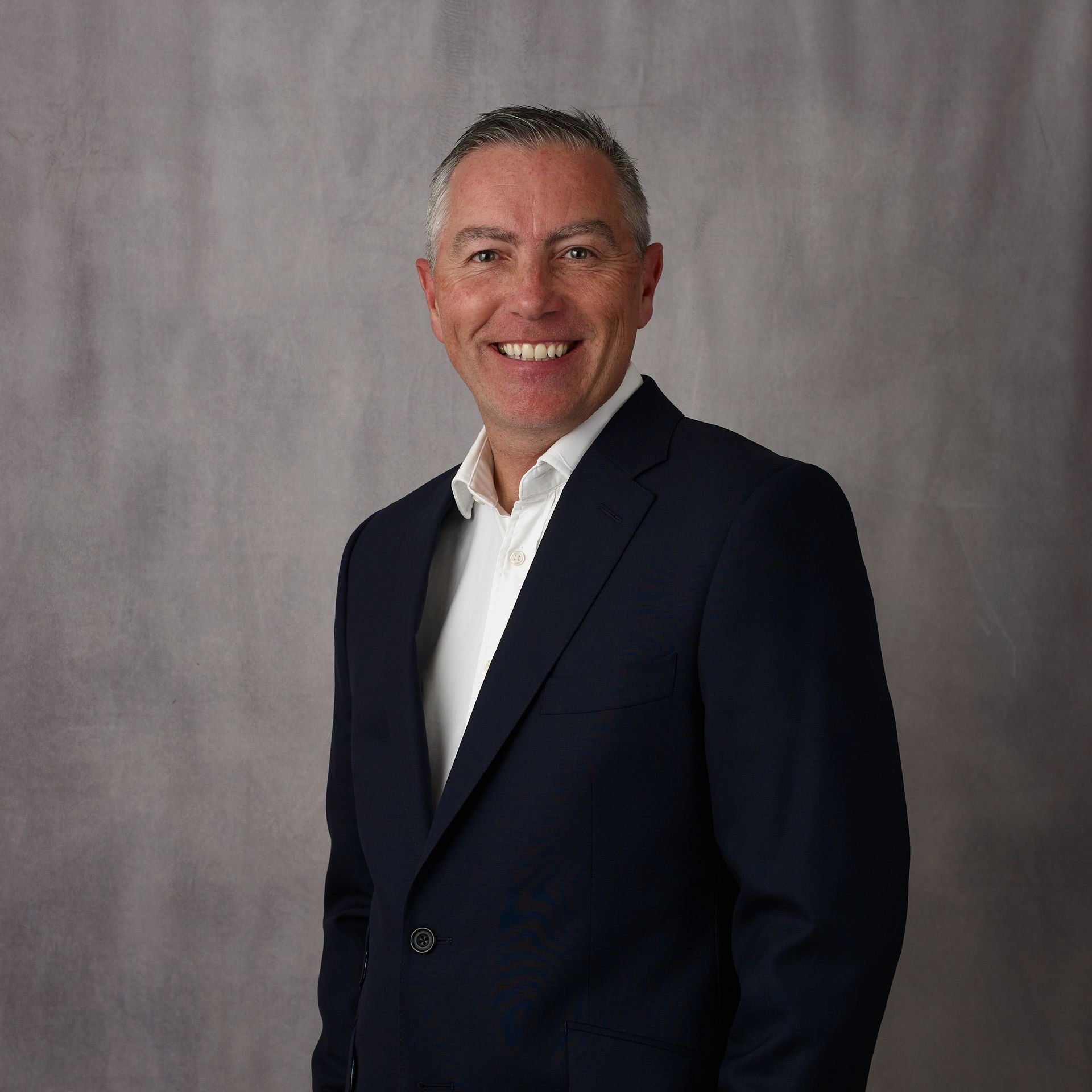 Man in dark suit smiles, posed against a gray backdrop.