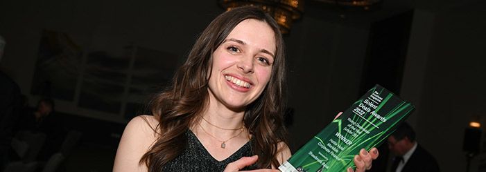 Woman smiling, holding a green award with text. Dark background, possibly an event.