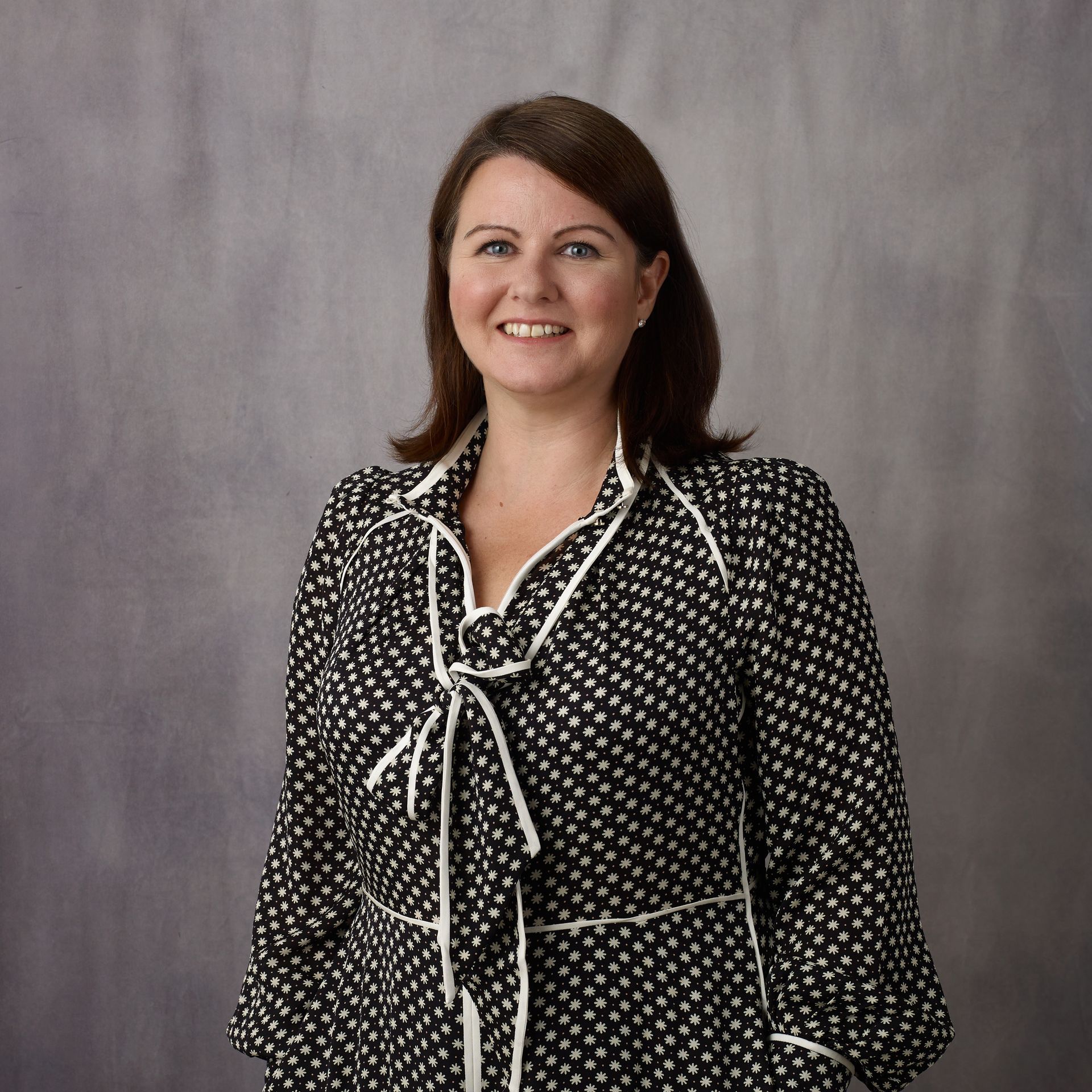 Woman with brown hair smiling, wearing a black and white polka dot blouse with a white trim, against a gray backdrop.