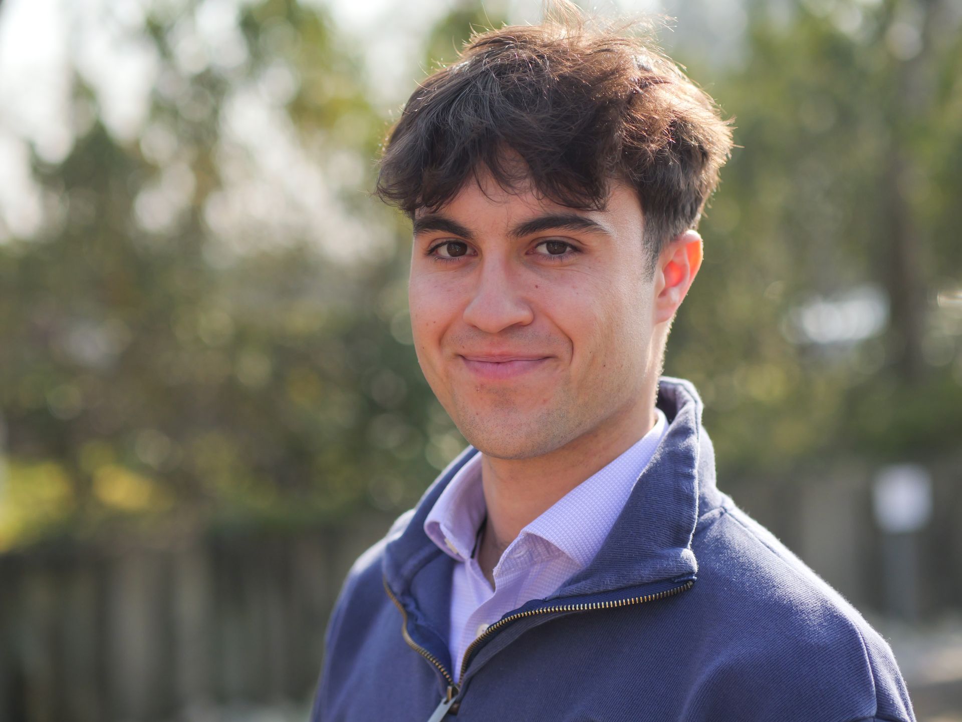 Man with short brown hair smiles, wearing a gray zip-up sweatshirt over a light blue collared shirt, outdoors.
