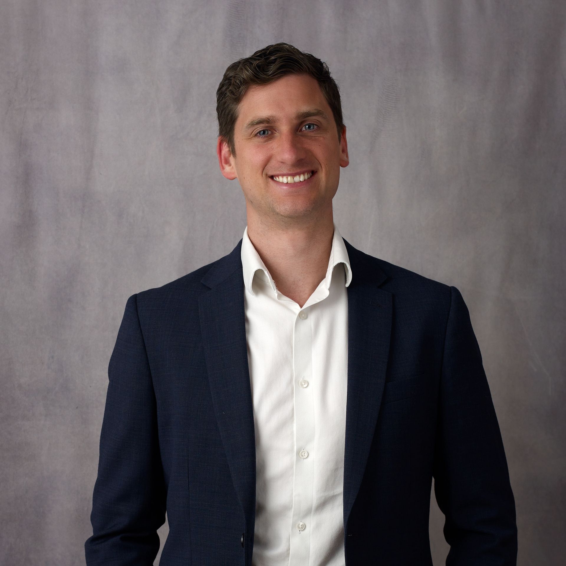 Man in dark blue blazer and white shirt, smiling, against a gray backdrop.
