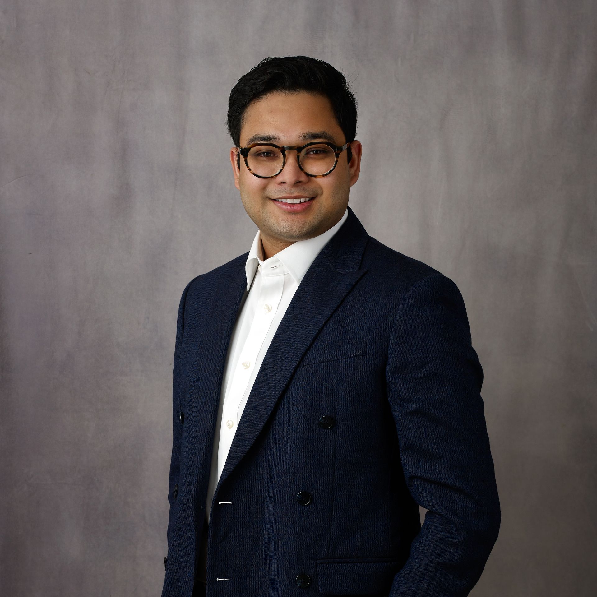 Man wearing glasses, white shirt, and navy blazer, smiling against a gray backdrop.
