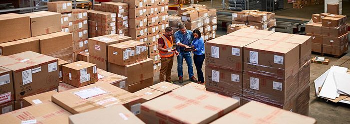 Three people looking at something in a warehouse, surrounded by stacks of cardboard boxes.