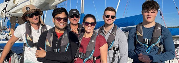 Group of people wearing life vests on a sailboat, posing with sunny sky in background.