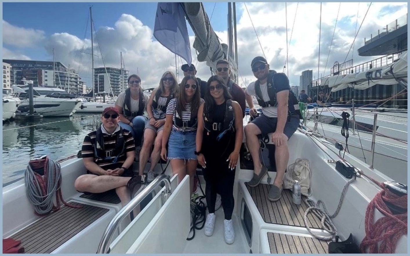 Group of people on a sailboat in a marina, smiling. Blue sky, boats, buildings visible.