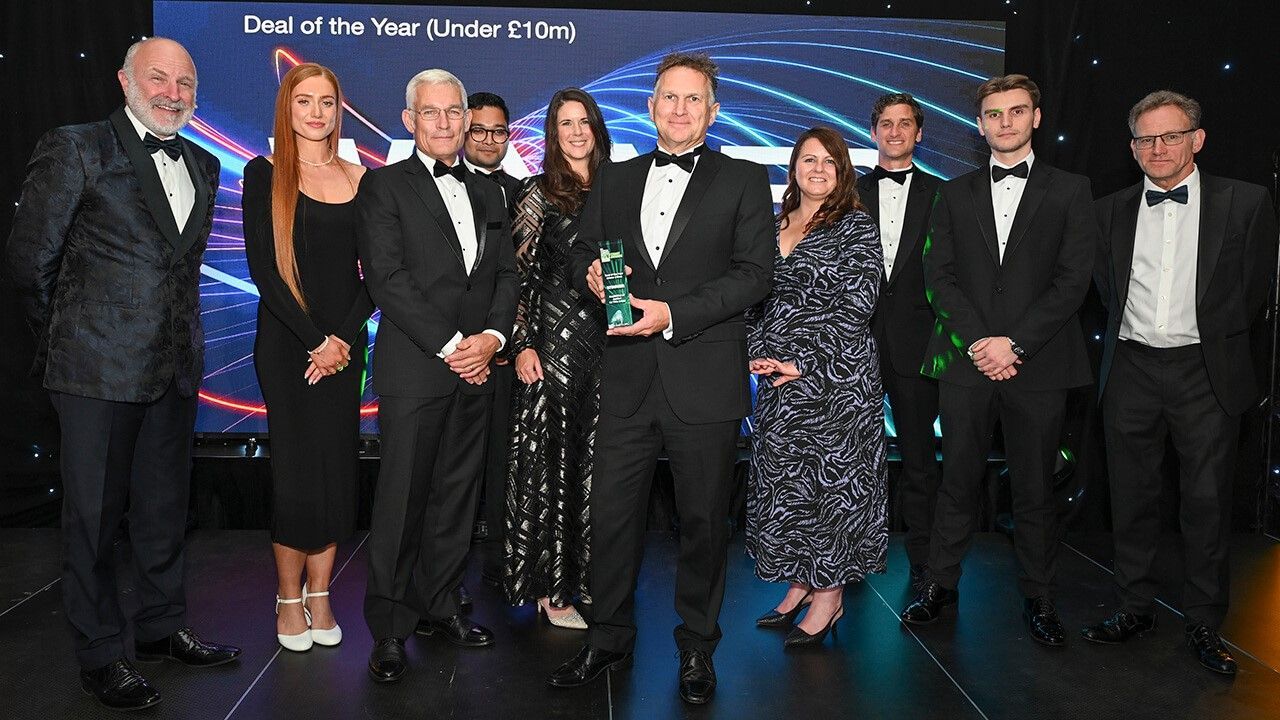 Group of people in formal wear on stage, some holding an award, at an event with a screen backdrop.