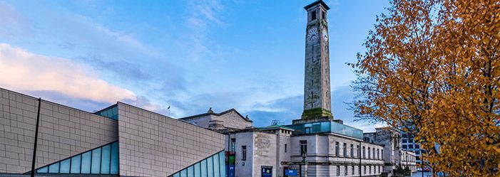 Tall clock tower next to a modern building, with autumn foliage in front and a blue sky above.