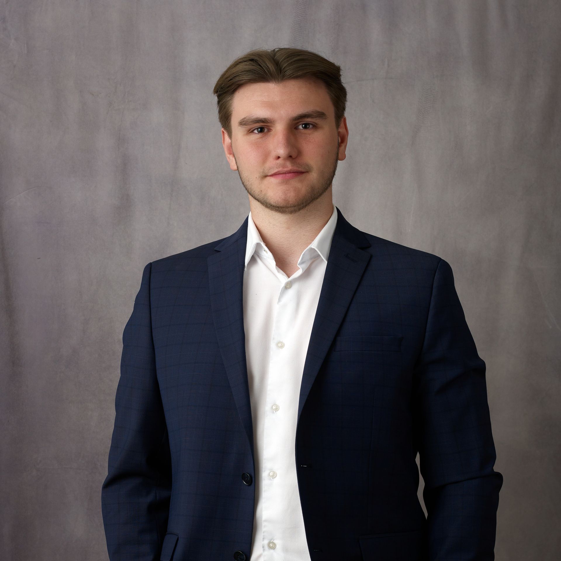 A young man with fair skin and short brown hair wears a dark blue suit and white dress shirt