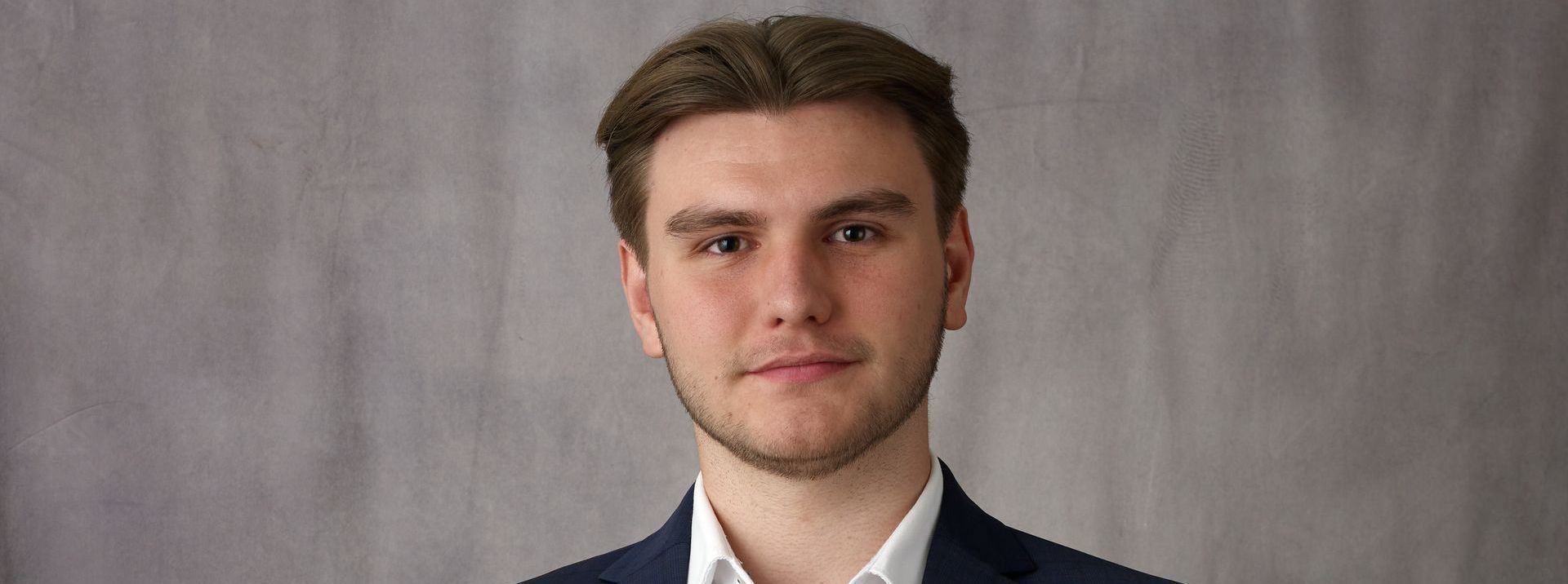 A man in a suit with a white shirt smiles at the camera against a neutral background.