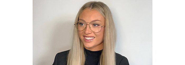Woman with long blonde hair and glasses smiles, wearing a black shirt in front of a white wall.