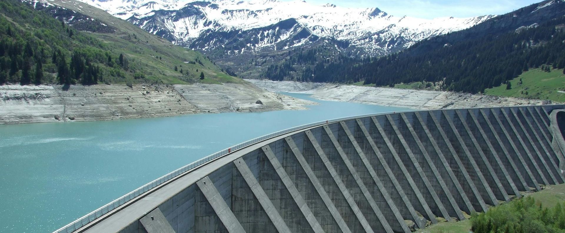 A concrete dam spans across a turquoise lake with snow-capped mountains in the background.