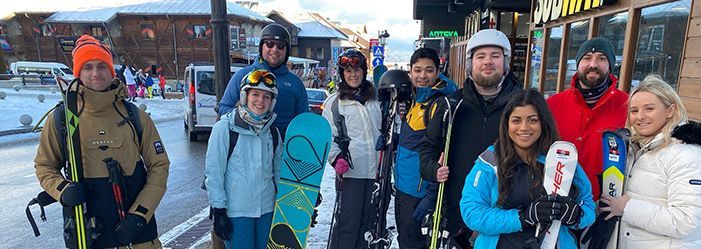 Group of people in ski gear posing outside near a snowy building, some holding skis or snowboards.