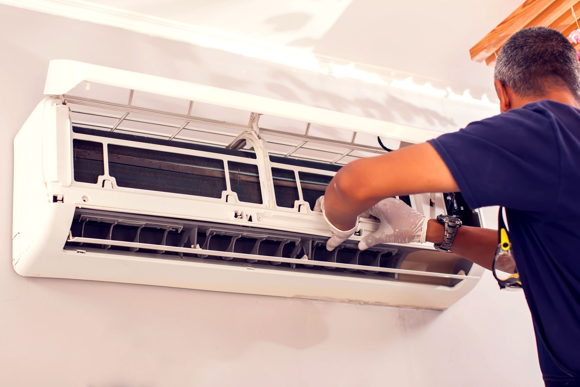 A technician wearing white gloves performs maintenance on a wall-mounted air conditioner.