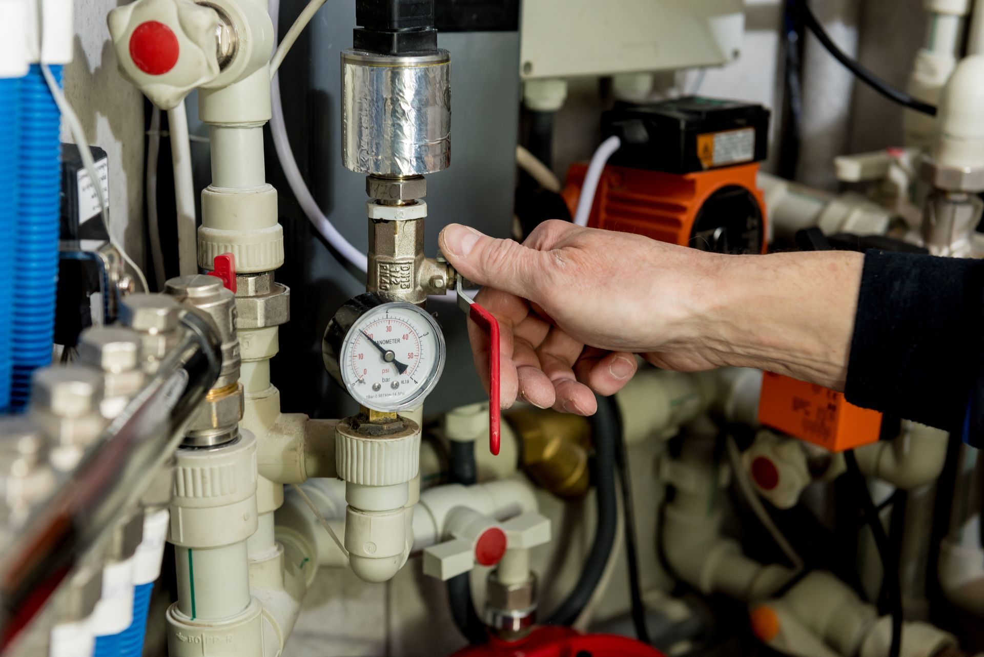 A hand turns a red valve lever on a residential boiler system near a circular pressure gauge.