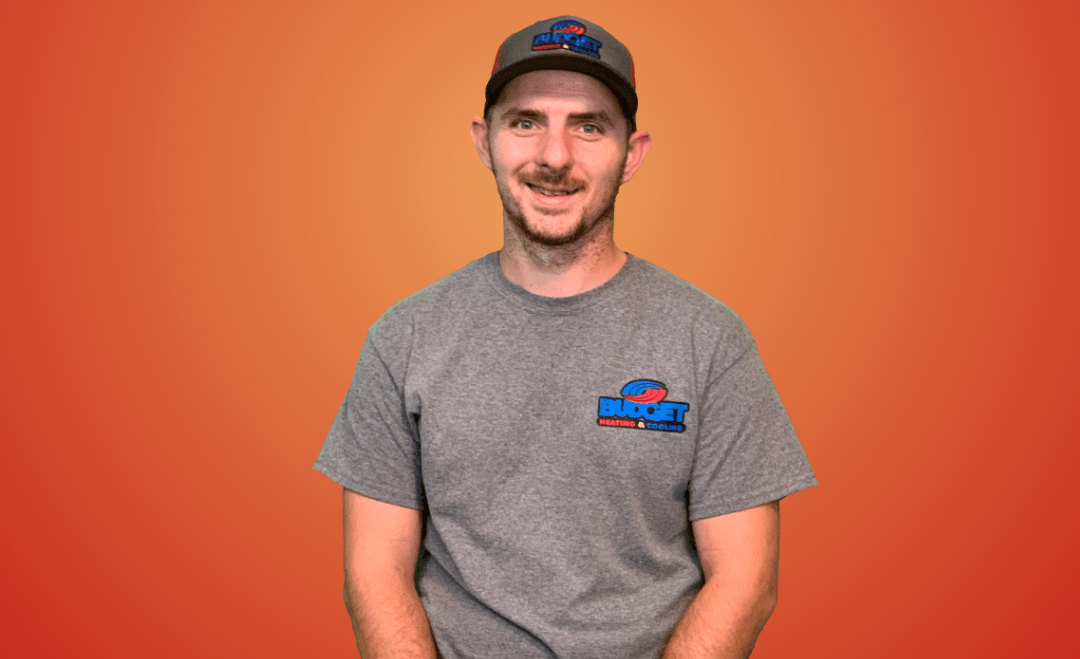 A smiling person wearing a grey shirt and cap with blue logos, posed against a solid orange background.