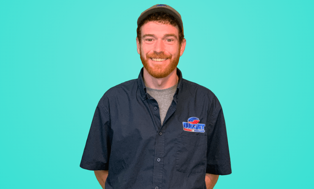 A person with red hair and a beard wearing a dark blue button-down shirt and a cap against a solid teal background.
