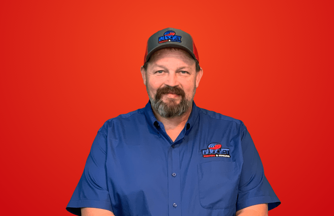 A person with a beard, wearing a blue shirt and a grey and red baseball cap, smiling against a solid red background.