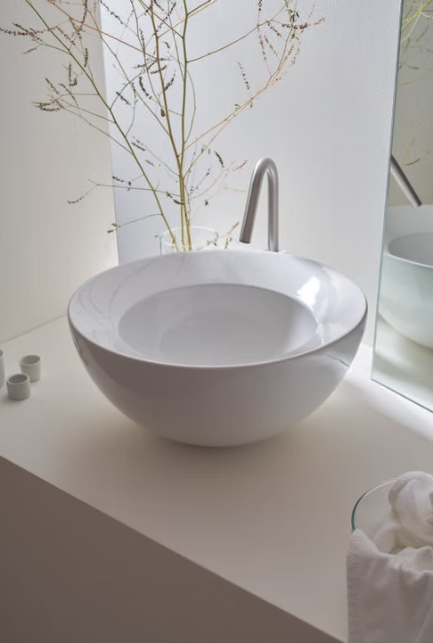 Lavabo moderno bianco a forma di ciotola su un piano di lavoro bianco accanto a una finestra con un vaso di rami spogli.