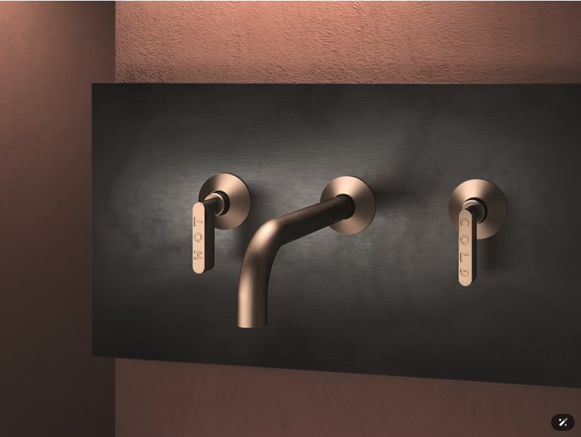Tre rubinetti moderni in bronzo montati su una parete scura sopra un lavabo.