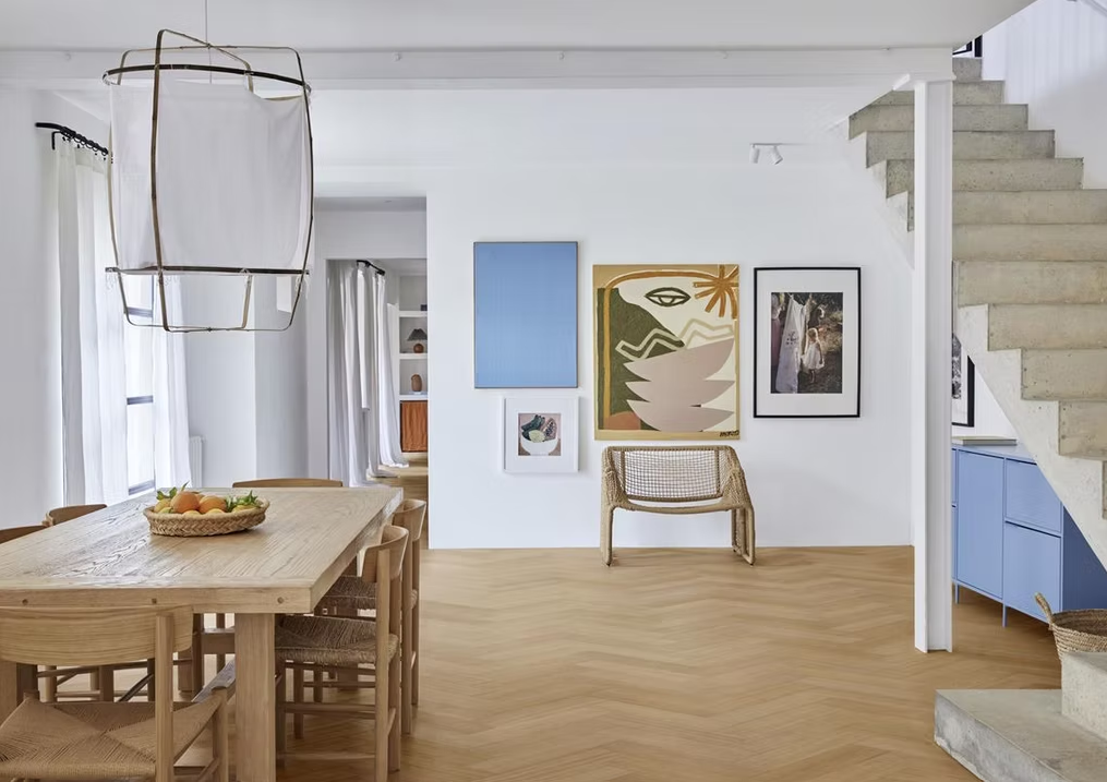 Sala da pranzo luminosa e minimalista con tavolo in legno, sedia in vimini, quadro blu alla parete e scala sulla destra.
