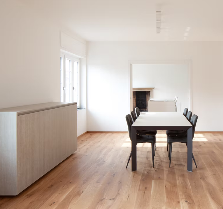 Sala da pranzo minimalista con tavolo in legno, sedie scure, credenza e ampie finestre in un luminoso ambiente bianco.
