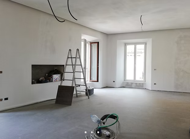 Stanza vuota parzialmente ristrutturata con scala, camino e finestre che lasciano entrare la luce naturale.