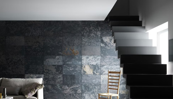 Stanza moderna con parete in pietra scura, scala nera, luce bianca dalla finestra e sedia in legno.