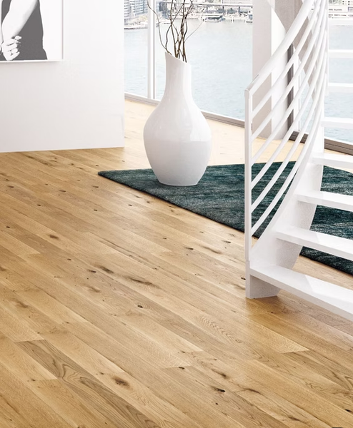 Luminosa stanza moderna con pavimento in legno chiaro, vaso bianco su tappeto scuro e scala bianca accanto a una finestra.