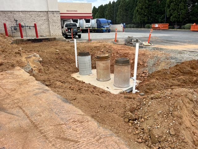 Construction trench with three utility pipes and markers beside a gravel lot and building