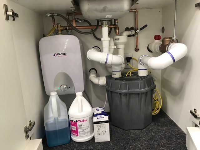 Under-sink plumbing with a water heater, cleaning supplies, and a gray bucket in a cabinet.