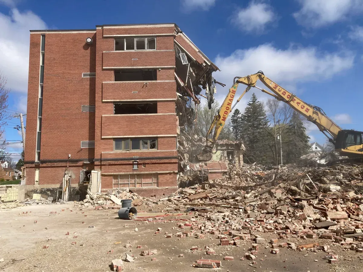A large brick building is being demolished by a yellow excavator with the word demolition on it