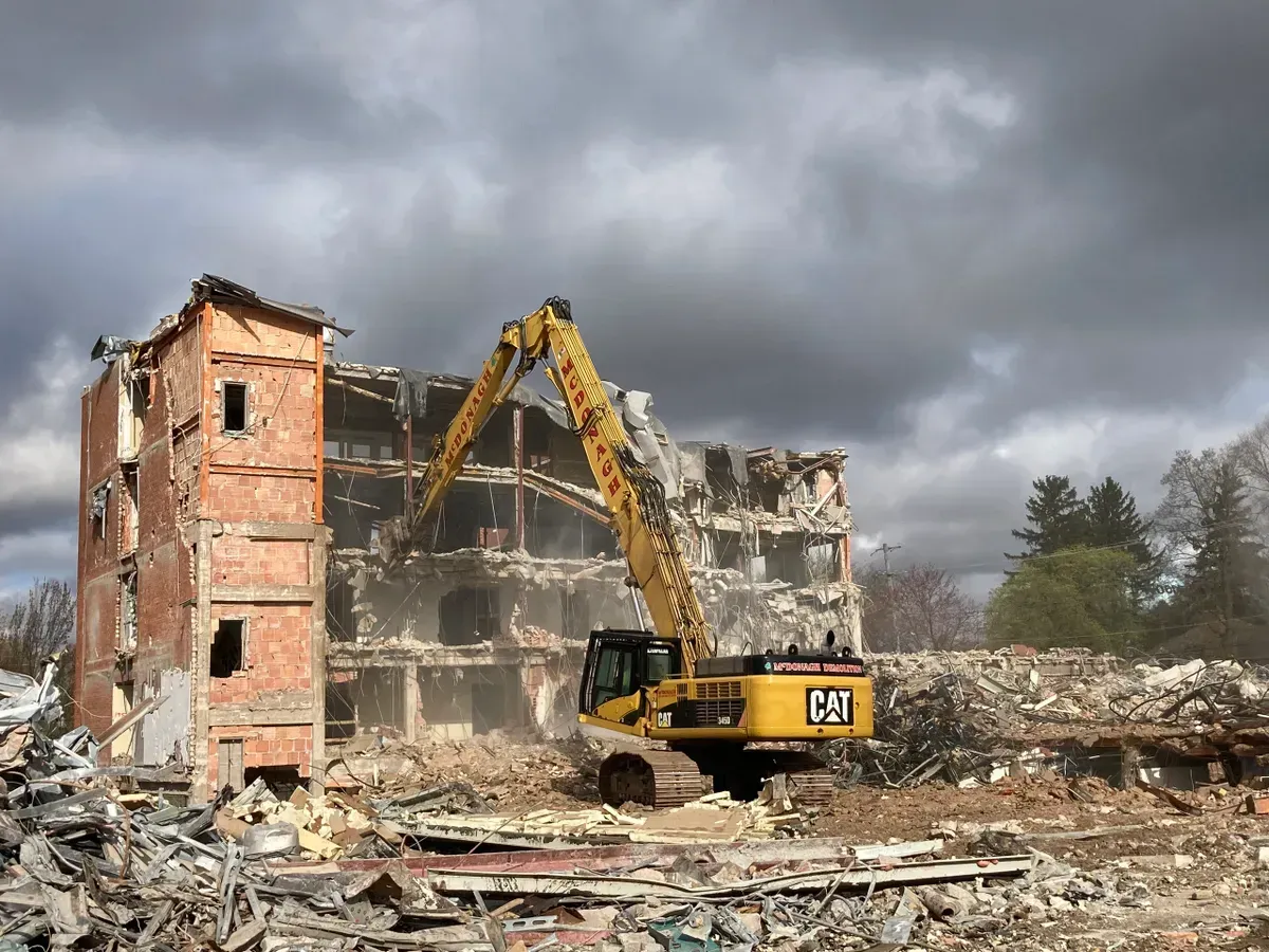 A large yellow excavator is demolishing a large brick building.