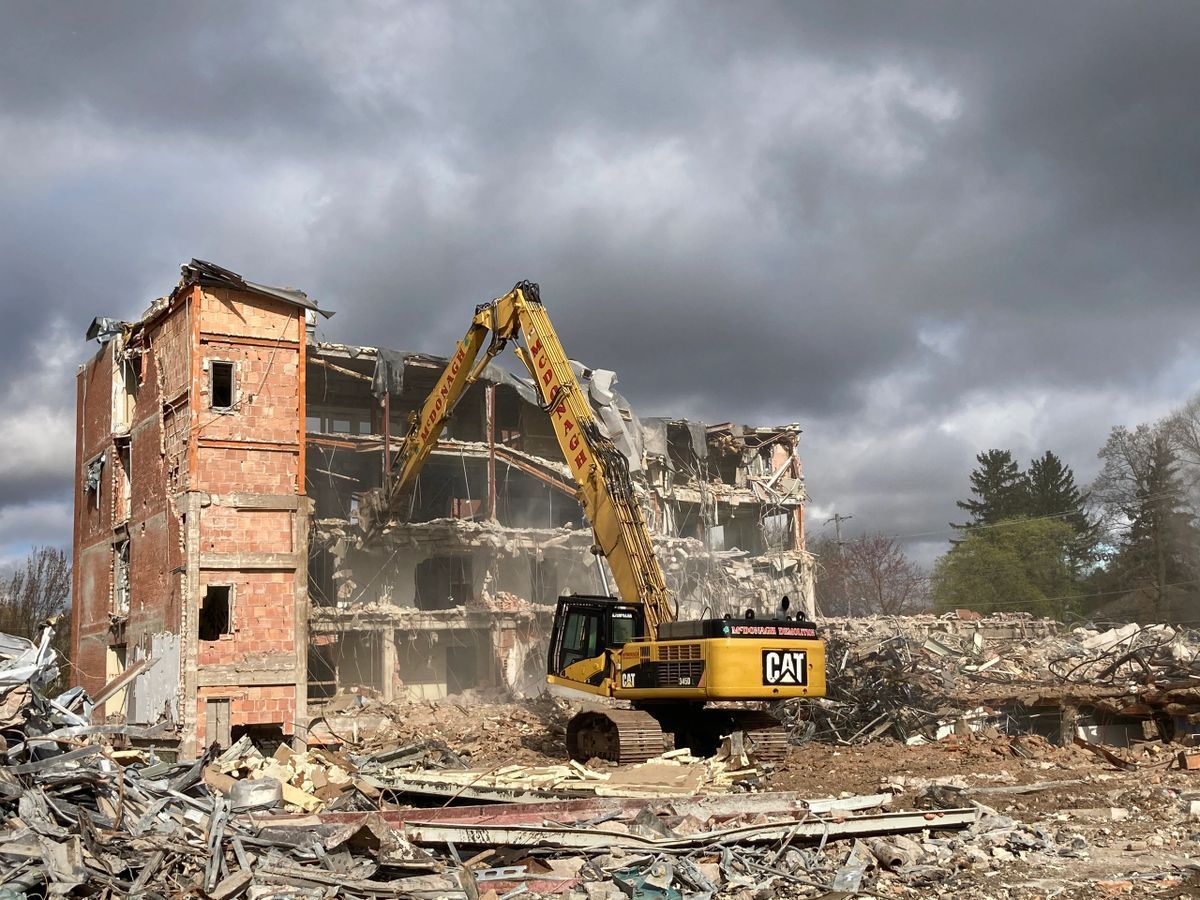 A large yellow excavator is demolishing a large brick building.