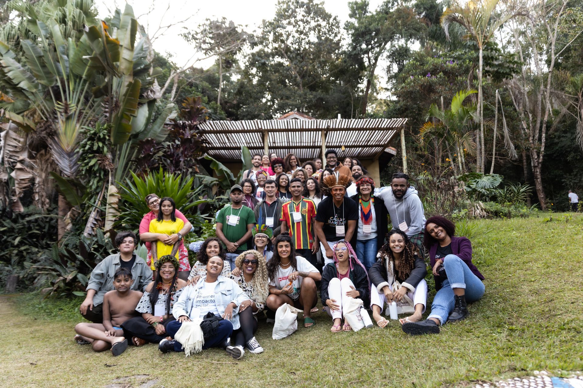 Um grande grupo de pessoas posa para uma foto ao ar livre em frente a uma estrutura rústica cercada por vegetação exuberante.