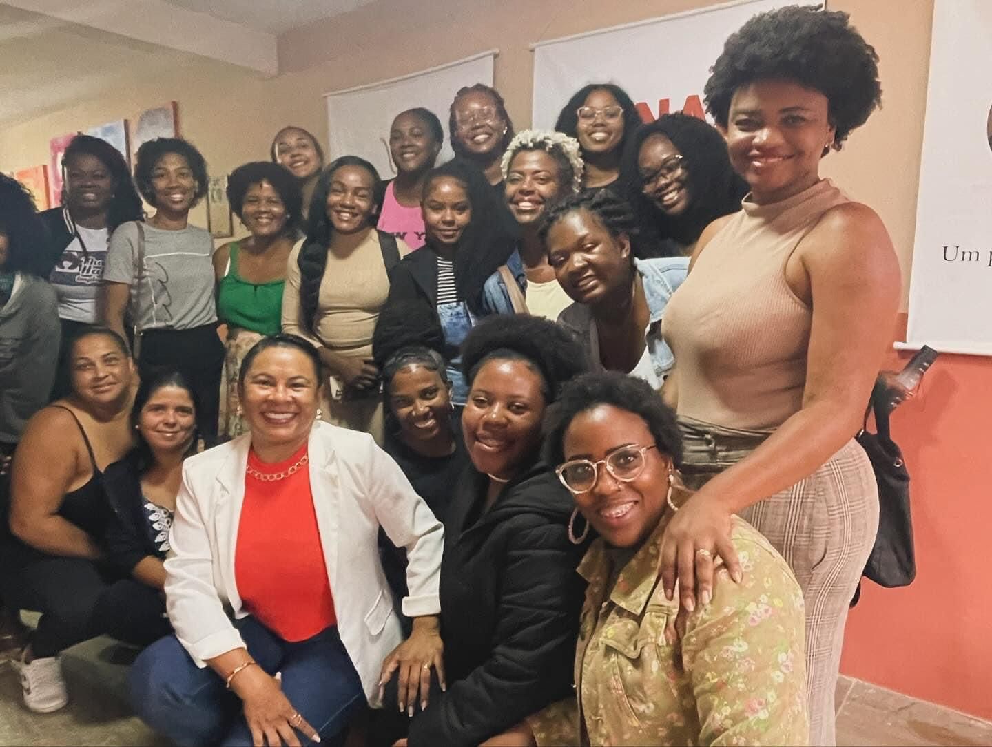 Um grande grupo de pessoas sorrindo e posando juntas em um ambiente interno, muitas vestindo roupas casuais de diversas cores.