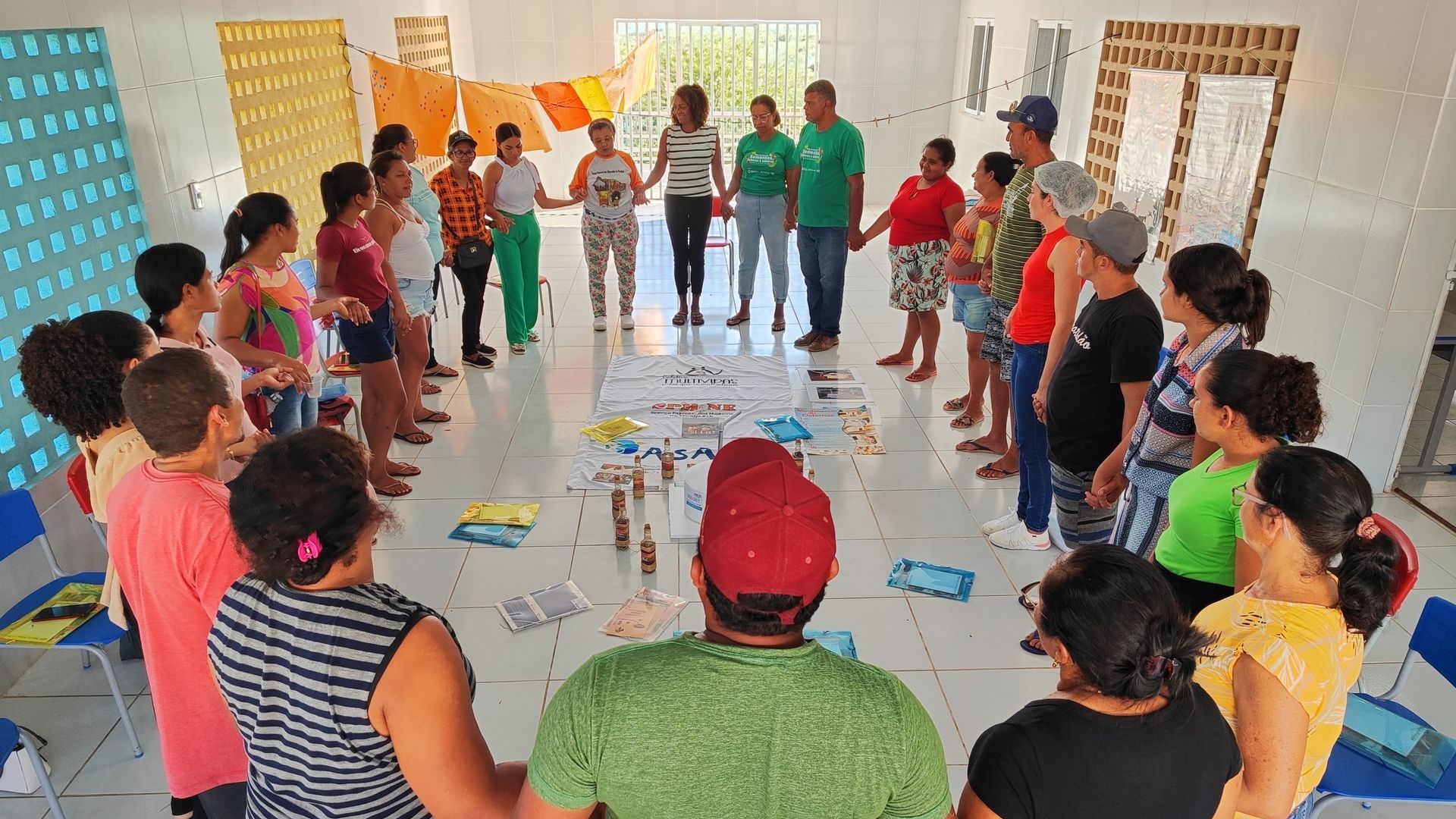Um grupo de pessoas está em um grande círculo em uma sala iluminada, de mãos dadas, para uma oficina ou atividade colaborativa.