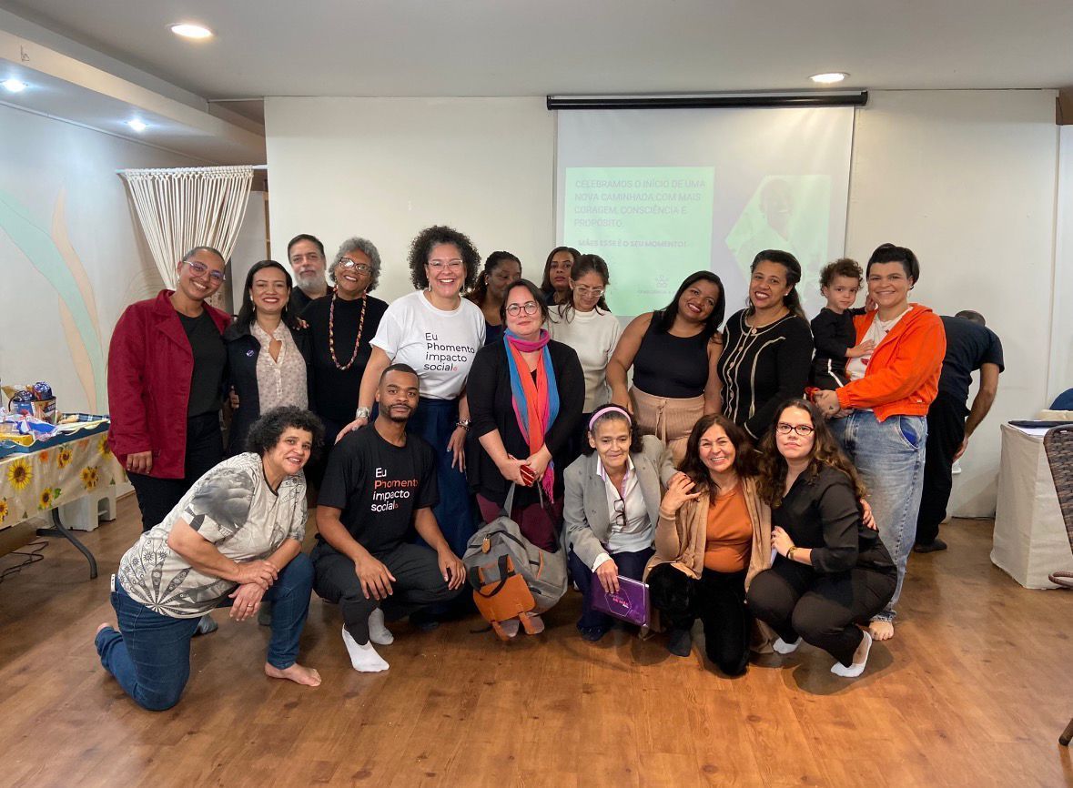 Um grupo de pessoas em pé, posando para fotos em uma sala com uma tela de projeção ao fundo, durante uma reunião ou workshop.