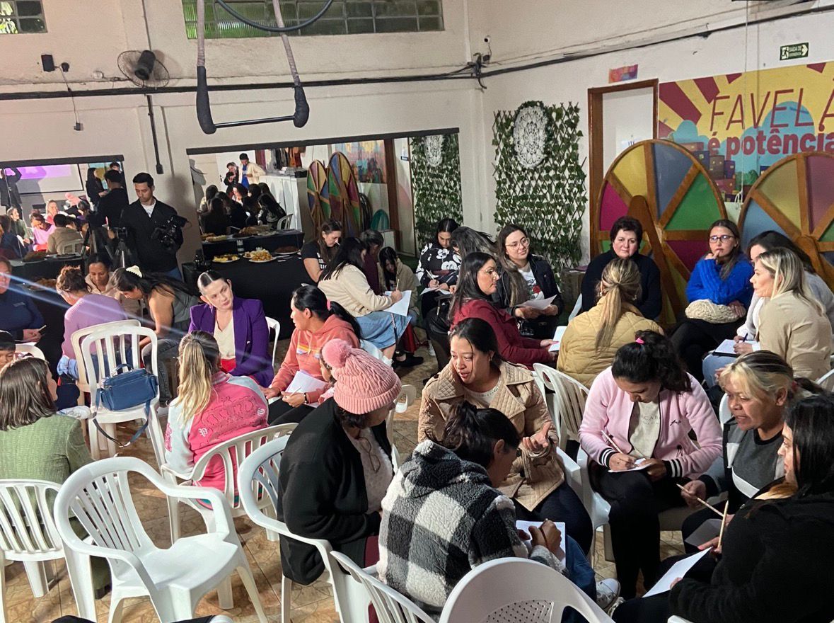Um grupo de pessoas senta-se em cadeiras dispostas em círculo para uma oficina ou reunião em um espaço interno com uma parede decorada.