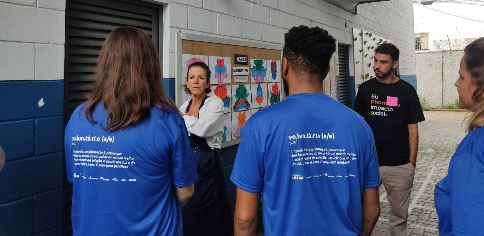 Um grupo de pessoas, algumas vestindo camisas azuis, está em uma sala com um quadro de avisos enquanto conversa com uma pessoa de avental.