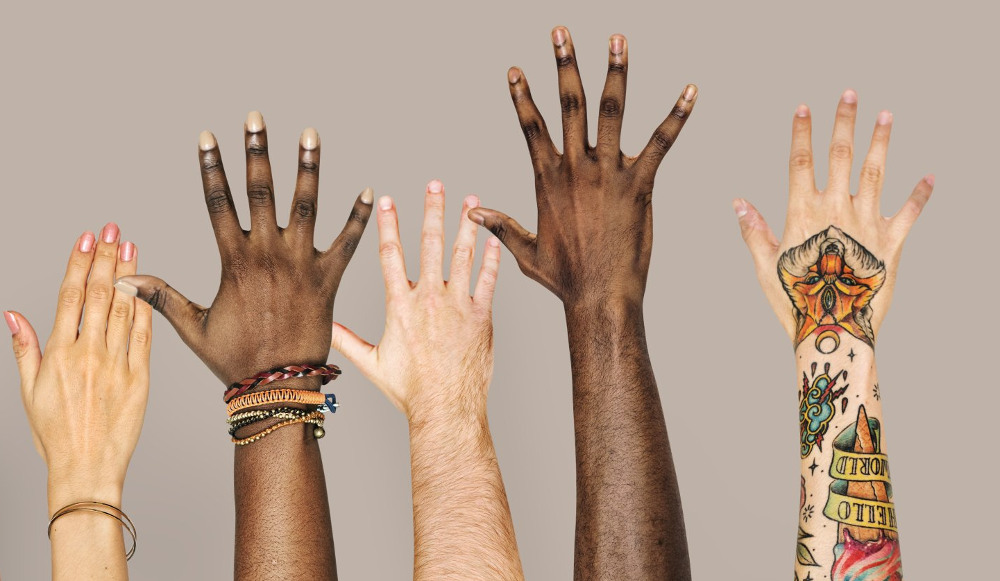 Cinco mãos diversas, com tons de pele variados, tatuagens e joias, erguidas contra um fundo claro e sólido.
