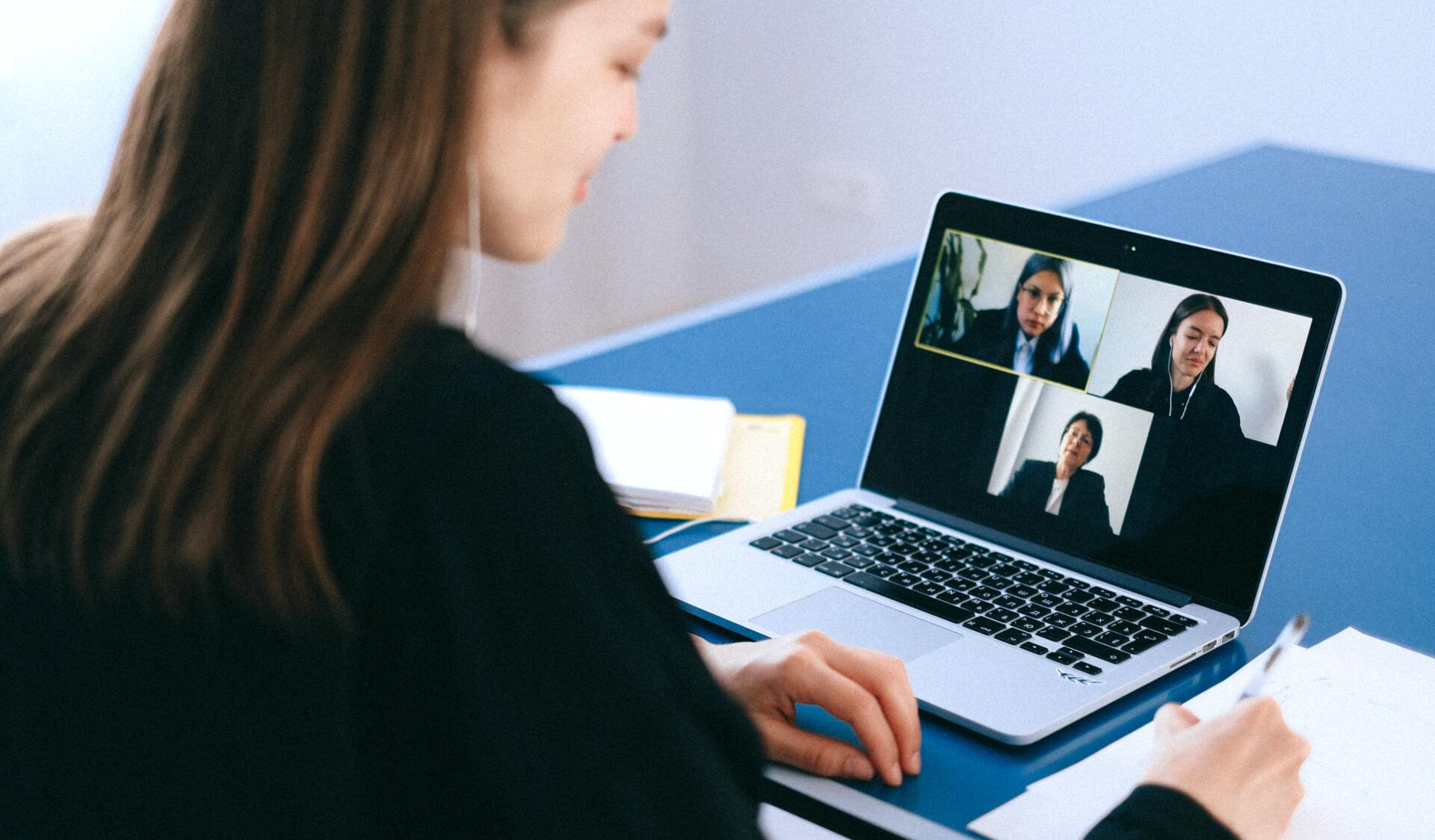 Uma pessoa está sentada à mesa, fazendo anotações enquanto assiste a uma videoconferência com outros três participantes na tela de um laptop.