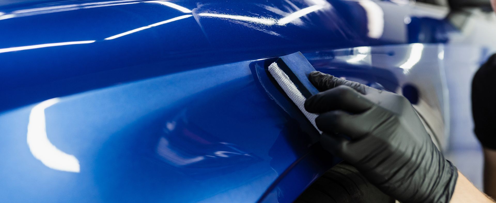 A person in black gloves is applying a detailing product to the blue paint of a car.