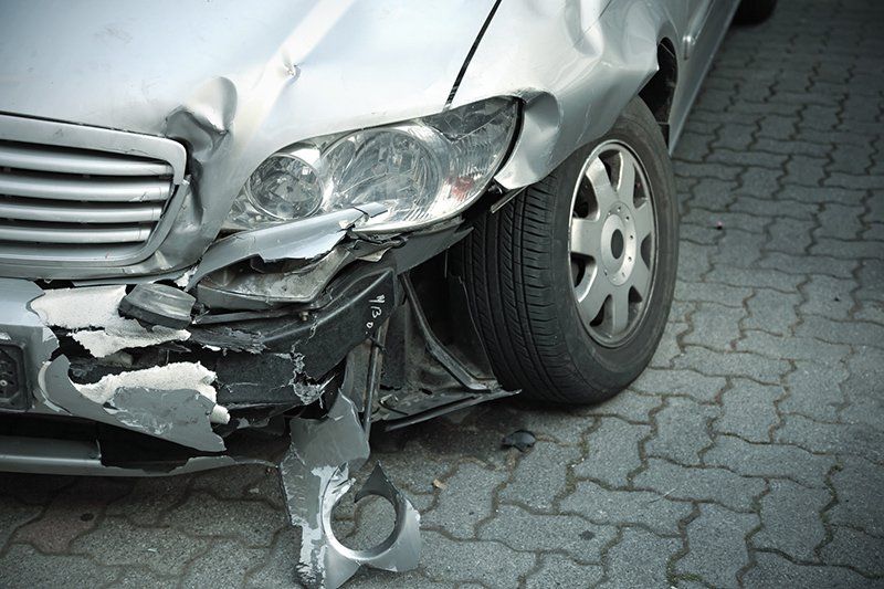 A silver car with a damaged bumper is parked on a brick sidewalk.