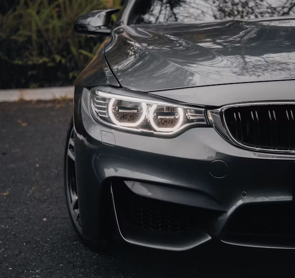 Front-left view of a gray sports sedan with headlights on, parked on a wet road in a dark wooded area