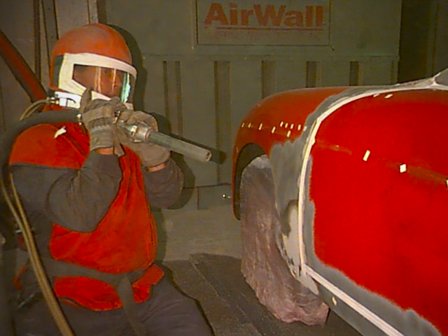 A man is sandblasting a red car in front of a sign that says airwall