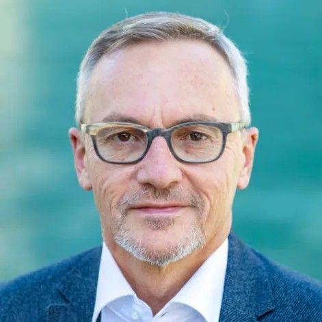 Porträt einer Person mit ergrauendem Haar und Brille, die ein weißes Hemd mit Kragen und einen blauen Blazer trägt, vor einem türkisfarbenen Hintergrund.