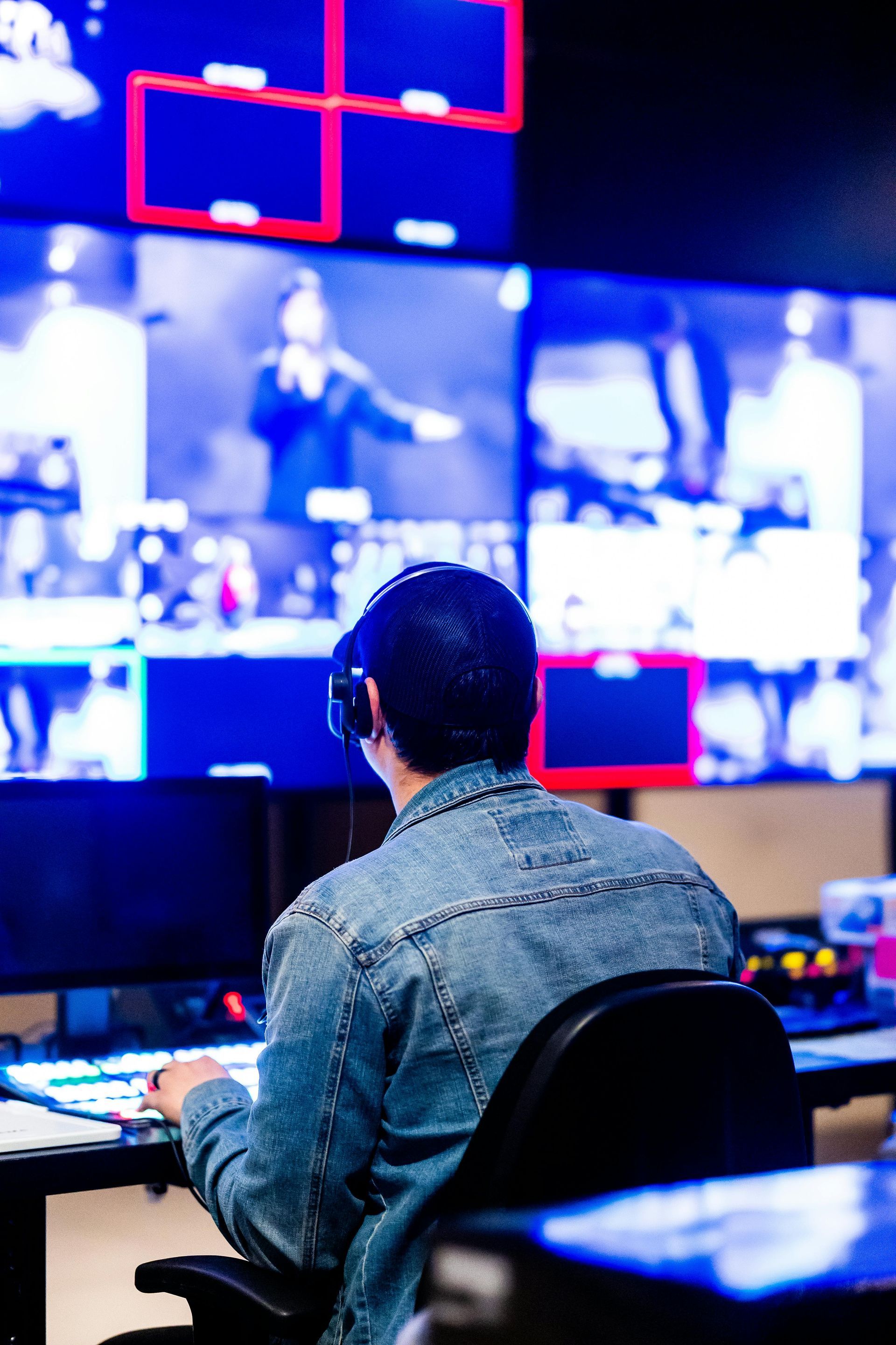 Person wearing headphones at a control panel, looking at multiple video screens in a dimly lit room.