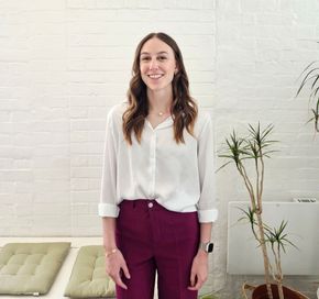 A picture of a smiling young women with long wavy brown hair against a white background with a couple of plants