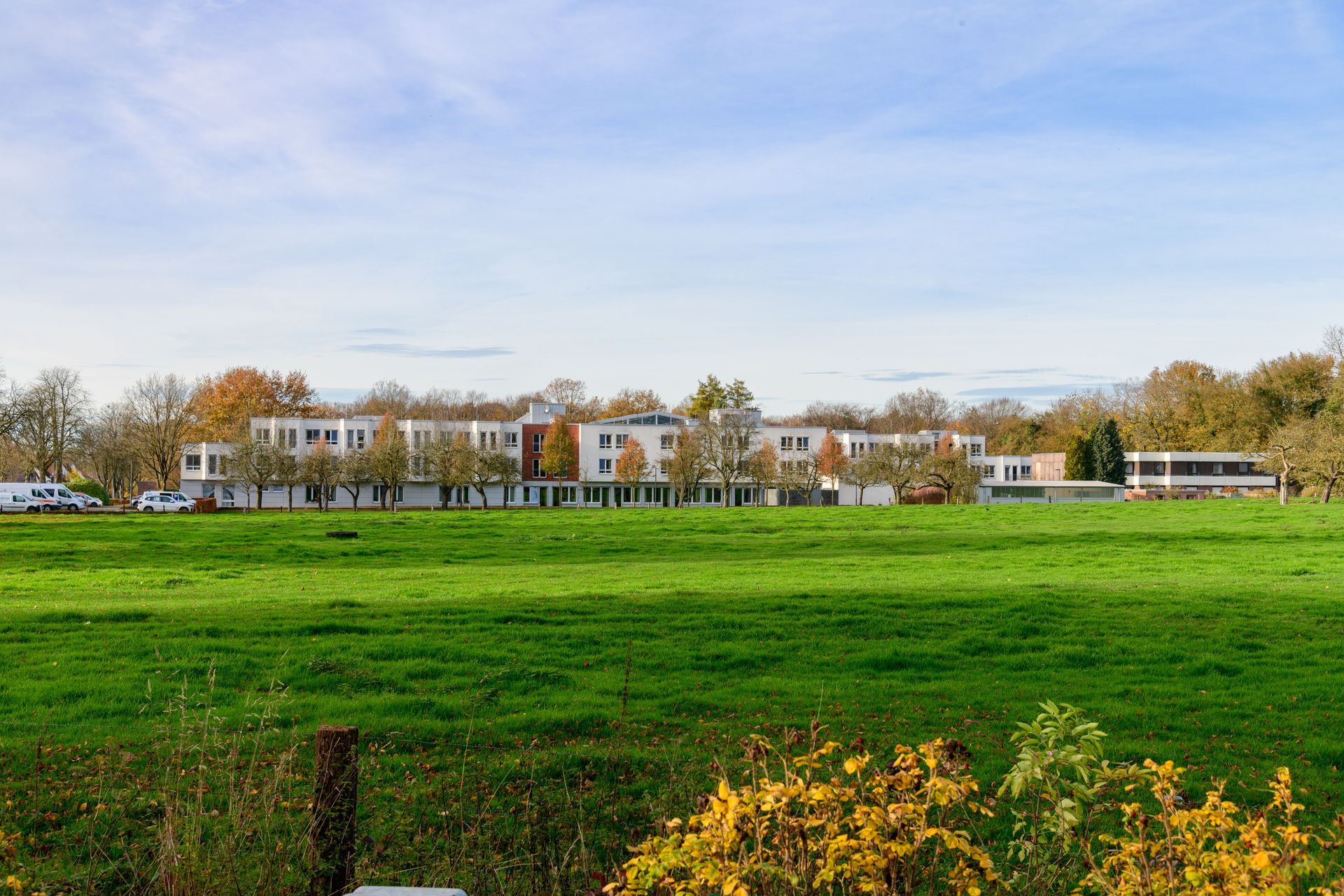 Een uitgestrekt groen veld met een lang wit gebouw en herfstbomen onder een blauwe hemel.
